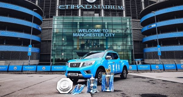Blue nissan navara parked in front of manchester city stadium, with 4 trophy cups