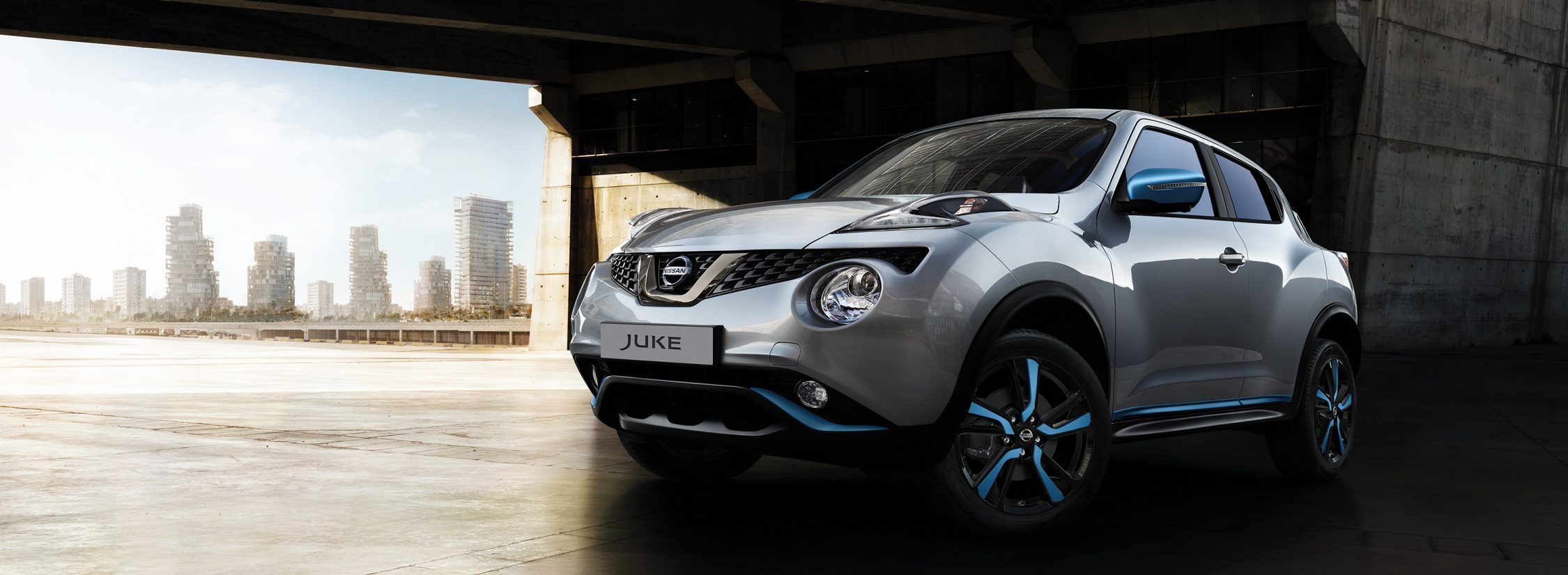 Nissan JUKE 2018: вид снизу спереди с поворотом на 3/4, автомобиль припаркован под мостом, элементы персонализации синего цвета (Power Blue)