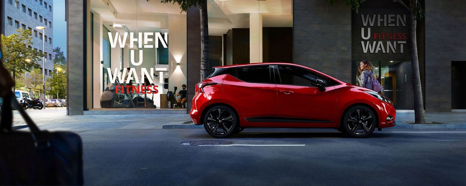 Profile shot of a red, New Nissan N-SPORT driving through the city with When U Want Fitness in the background