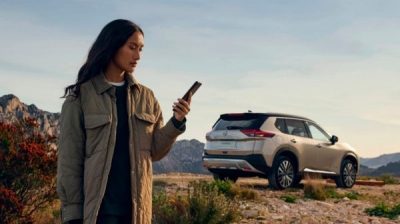 Eine Frau sieht neben einer 3/4-Heckaufnahme des X-Trail in den Bergen auf ihr Smartphone