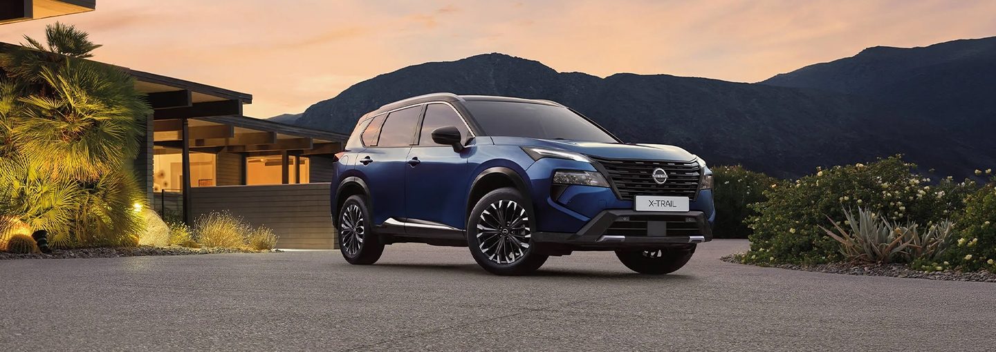 Plavi Nissan X-Trail - prednji bočni pogled ispred kuće pri zalasku sunca