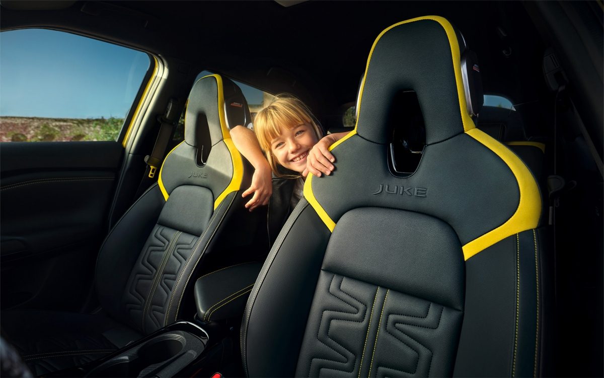 Ein Mädchen blickt zwischen den Vordersitzen eines Nissan Juke mit schwarzem Leder und gelben Akzenten in die Kamera.