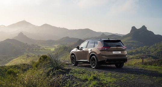 Nissan X-Trail in Dunkelbeige mit schwarzem Dach von hinten auf einem Feldweg in einer sommerlichen Hügellandschaft