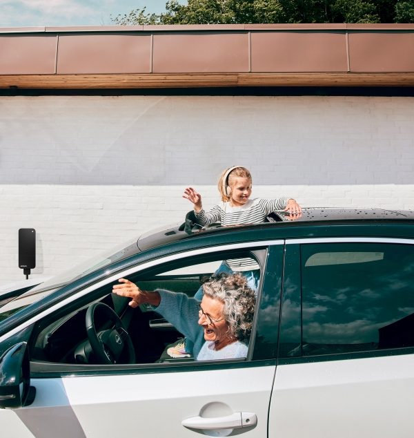 En ældre kvinde bag rattet i en hvid Nissan elbil smiler og vinker ud af vinduet, mens et barn kigger ud af soltaget. I baggrunden er en sort Norlys ladeboks monteret på en hvid mur.