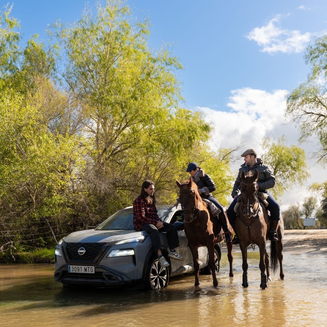 Un vehículo Nissan detenido a la orilla de un río con Lethal sentado en el el capó del mismo mientras conversa con unos señores que están sentados en sus caballos.