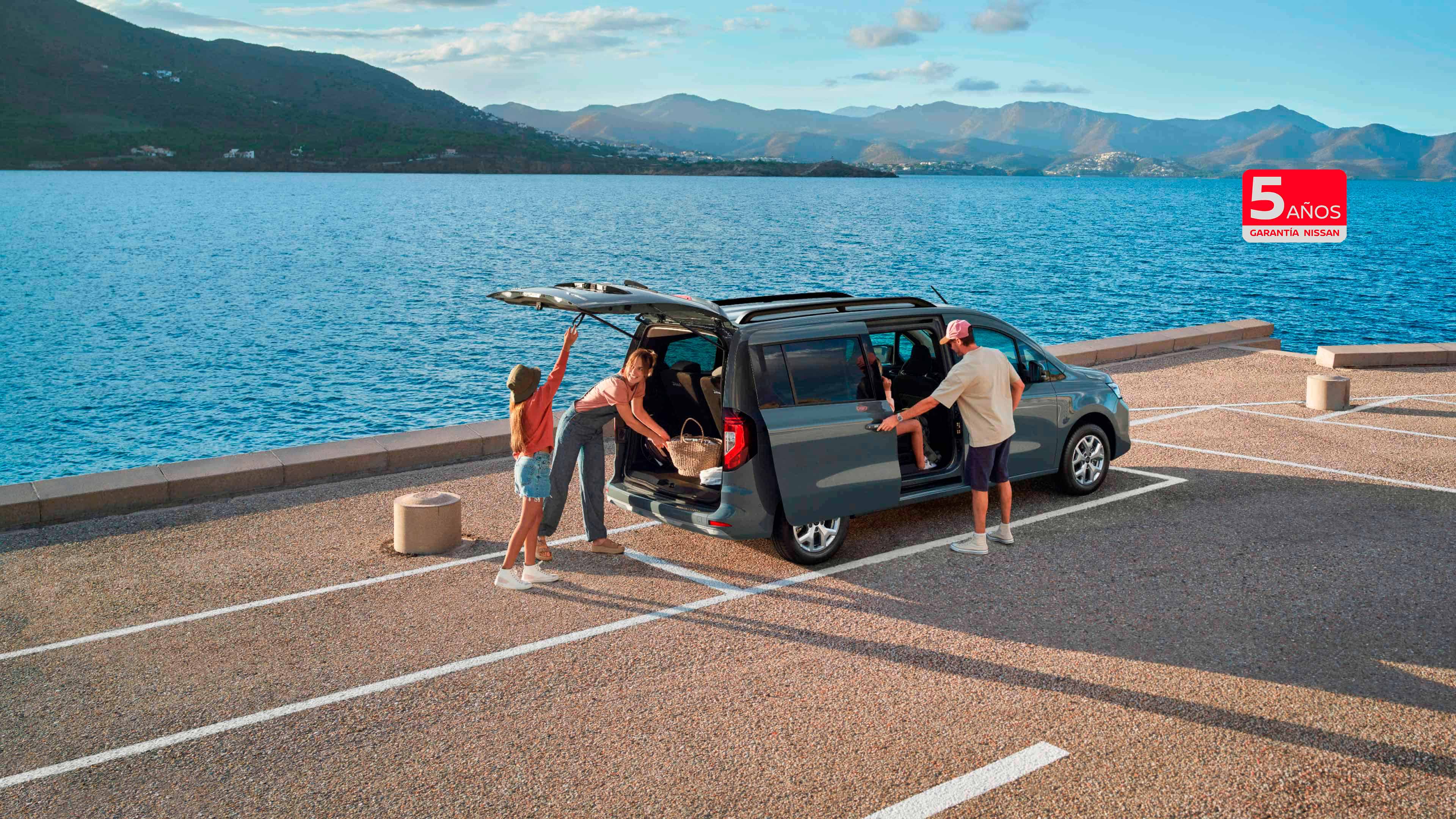 Nissan TOWNSTAR Combi: vista frontal de 3/4 de la Combi aparcada en la playa con una pareja sujetando unas tablas de surf y mirando al mar al atardecer