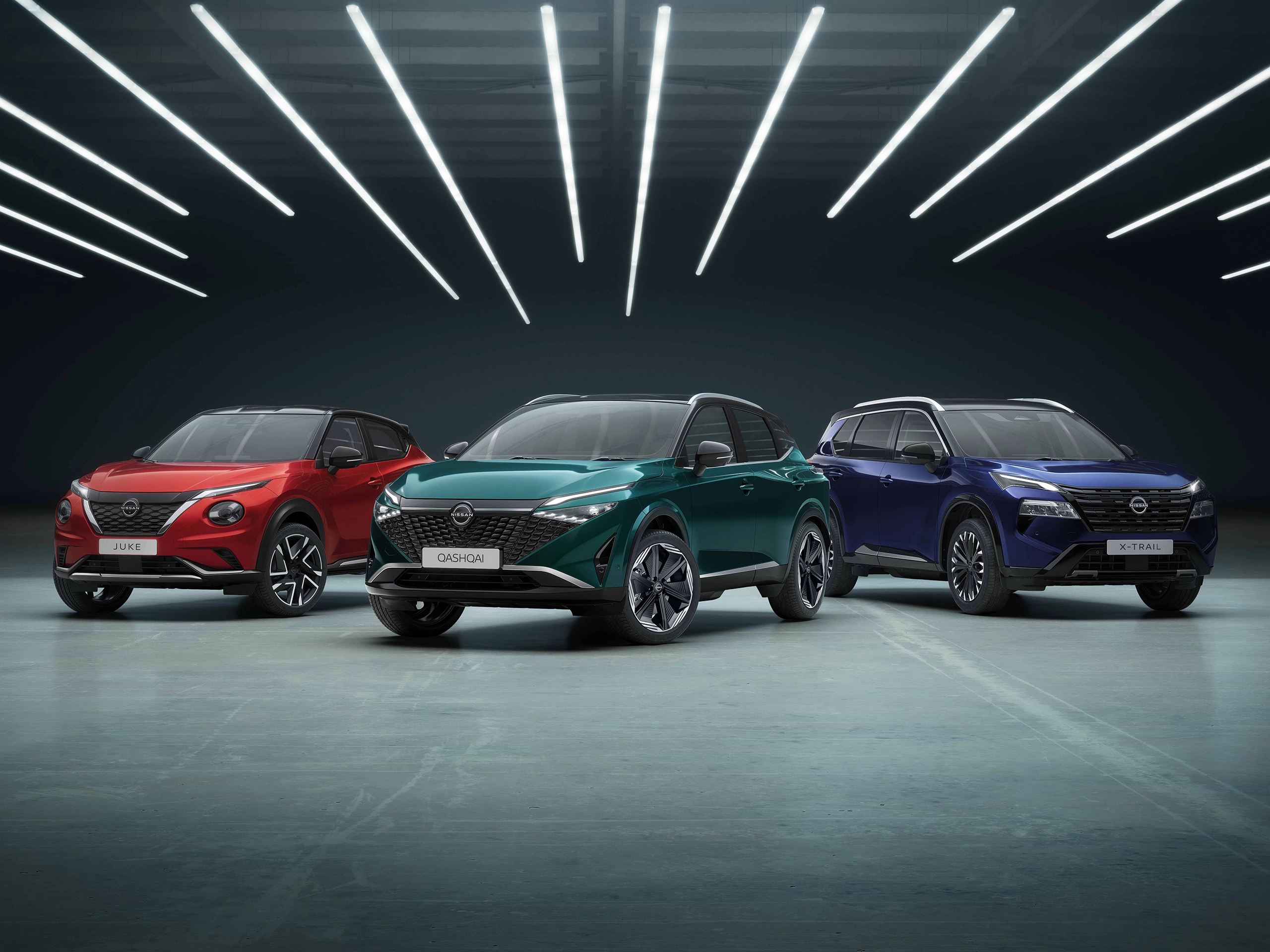 A trio of modern Nissan SUVs parked side-by-side in a sleek, dimly lit studio under striking, linear overhead LED strip lights. The lineup includes a red Nissan Juke on the left, a teal-green Nissan Qashqai in the centre, and a blue Nissan X-Trail on the right, all showcasing their front designs on a polished concrete floor.