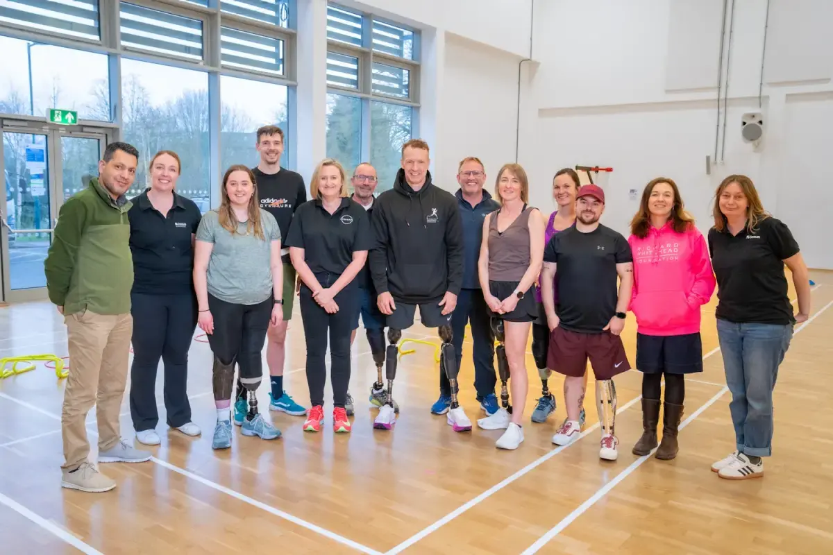 Group of people, including Richard Whitehead in the centre, standing side by side in a sports hall, several wearing athletic clothing and prosthetic lower limbs, with training equipment visible in the background.