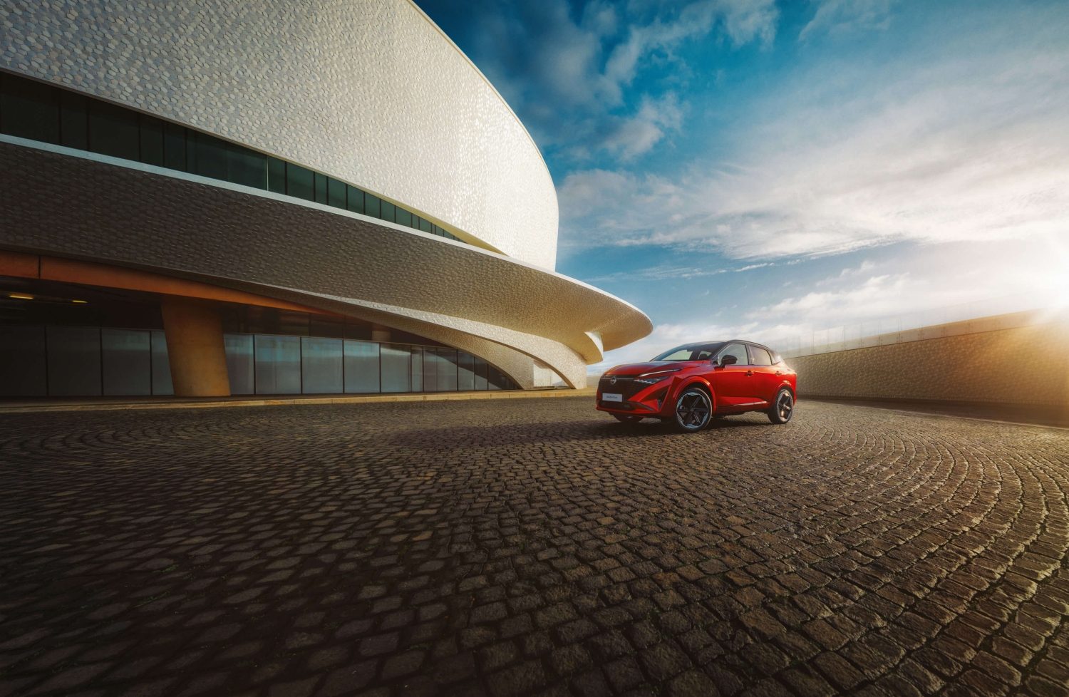Nissan Qashqai e-POWER 3/4 priekšpuses skats, novetots pie ēkas, un tam tuvojas sieviete – SUV hibrīds.