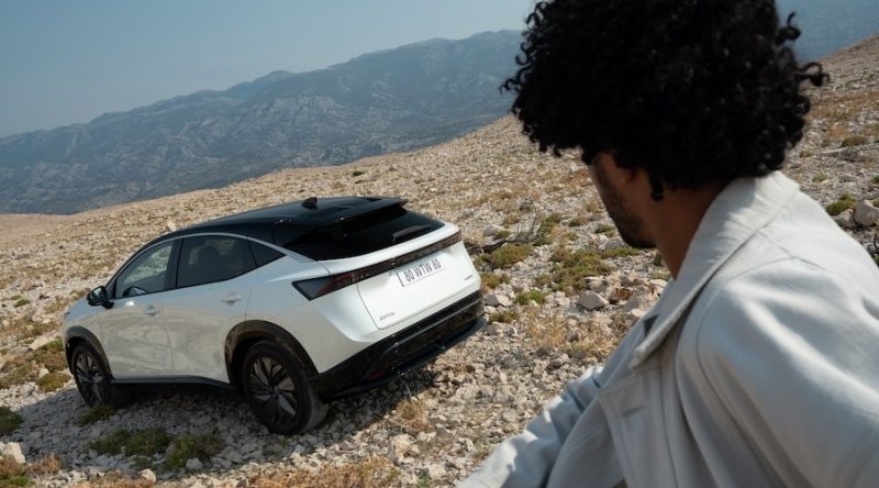 an met krullend haar kijkt uit op ARIYA bovenop een heuvelmet Nissan-logo op de voorgrond